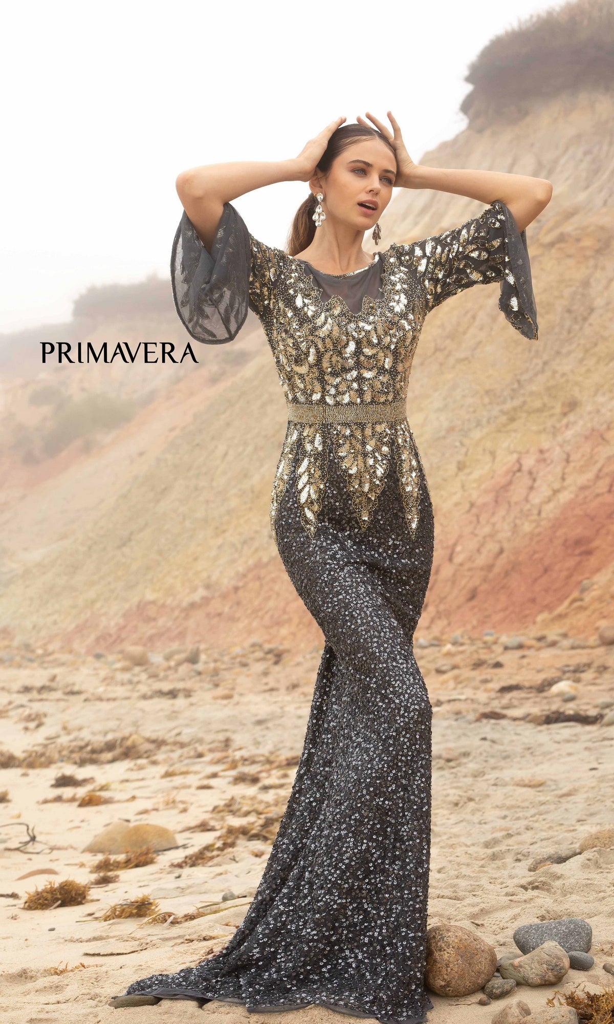A woman in the Bell-Sleeve Long Beaded Evening Gown 1424 poses on a rocky beach with cliffs in the background.